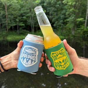 May include: Two beverage insulators, one light blue with a UFO graphic and the word "UFOLOGIST", the other green with a Cryptid Ranger design. A can and a bottle are held in the insulators, with a forest and water background.