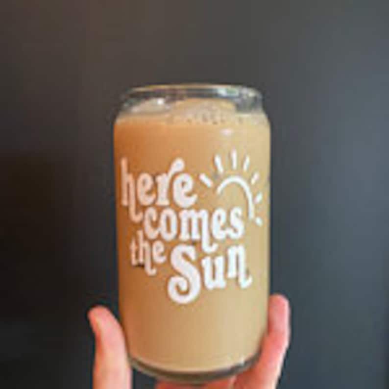 May include: A glass can with the text "here comes the sun" and a sun graphic. The can is filled with a light brown beverage.