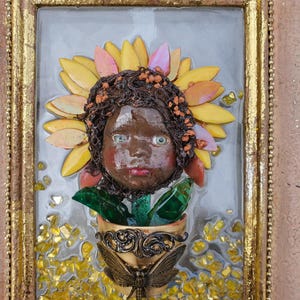 May include: A framed artwork featuring a doll's face with a sunflower headdress. The doll has brown skin, green eyes, and red lips. A decorative pot with a dragonfly is at the bottom. The frame is gold and the background is gray.
