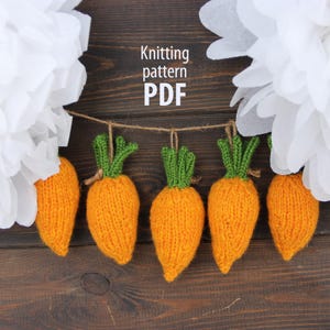 May include: A string of five knitted orange carrots with green tops hangs against a dark wood background. The text "Knitting pattern PDF" is visible. White paper flowers are in the background.