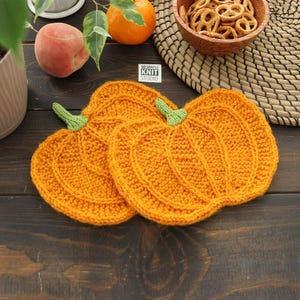 May include: Two orange pumpkin-shaped knit coasters with green stems. The coasters feature a textured ribbed pattern and are positioned on a dark wooden surface. A small white tag with the text SO SIMPLE KNIT STUDIO is visible between the items.