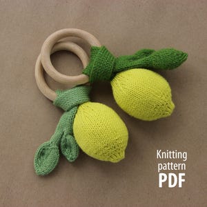 May include: A handmade baby rattle featuring two wooden rings and two knitted lemons with green leaves. The lemons are yellow, and the leaves are green. The text "Knitting pattern PDF" is visible in the lower right corner.