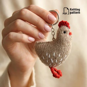 May include: A knitted chicken keychain in shades of tan, red, and white. The chicken has a red comb and feet, with white stitching details. The keychain includes a metal ring and chain. Text on the image reads "Knitting pattern PDF".