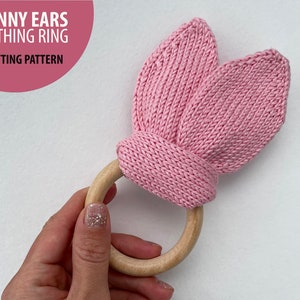 May include: Pink knitted bunny ear teether with a wooden ring.  The knitted ears are attached to the ring.  "Bunny Ears Teething Ring Knitting Pattern" is written on the image.