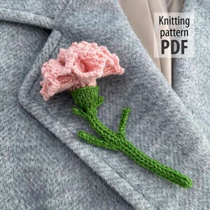 Puede incluir: Una flor rosa tejida a mano con un tallo y hojas verdes está prendida a un abrigo gris. La flor tiene pétalos texturizados. El texto "Knitting pattern PDF" está en la esquina superior derecha.