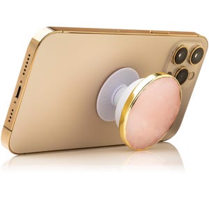 May include: A gold smartphone with a collapsible phone grip attached to the back. The grip features a circular, pale pink stone face with a metallic gold-colored edge. The base of the grip is white and displays the word CRYSTALFY in embossed letters.