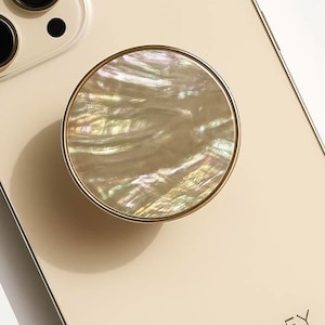 May include: A round phone grip with a gold-colored rim and a pearlescent, iridescent center. The phone grip is attached to a cream-colored phone case. The word "CRYSTALFY" is printed on the phone case.