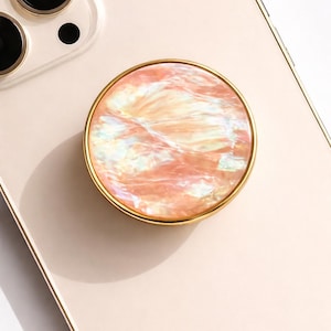 May include: A round phone grip with a pearlescent, peach-colored surface and a gold-colored metal rim. The phone grip is attached to a light pink phone case. The word "CRYSTALFY" is printed in gold on the phone case.