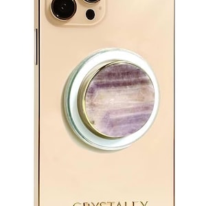 May include: A gold-colored smartphone with a triple-lens camera system and the text CRYSTALFY printed at the bottom. Attached to the center is a circular phone grip featuring a polished, banded purple and white stone inlay with a gold rim.