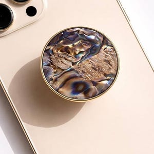 May include: A round phone grip with a gold-colored rim and a swirling, iridescent abalone shell design. The phone grip is attached to a light beige phone case. The word "CRYSTALFY" is printed on the phone case.
