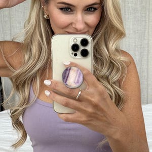May include: A light purple tank top paired with matching shorts. The phone held in the center features a circular grip attachment with a purple and white marbled pattern, showcasing a triple-lens camera system on the back of the device.
