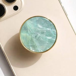 May include: A round phone grip with a gold-colored rim and a light blue and white marbled design. The phone accessory is attached to a cream-colored phone case. The word "CRYSTALFY" is printed on the phone case.