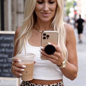 May include: A gold smartphone with a black circular grip attached to the back, a white sleeveless crop top, and high-waisted pants featuring a brown and black leopard print pattern. The outfit includes a gold necklace, a gold wristwatch, and rings.