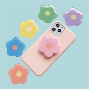 May include: A pink phone case with a flower-shaped phone grip attached. Six additional flower-shaped phone grips in various colors, including yellow, blue, green, purple, and orange, are scattered around the phone. The grips have a light-colored center.