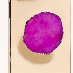 May include: A gold-colored phone case with a vibrant, fuchsia-colored agate stone inlay. The case features the word "CRYSTALFY" in gold lettering. The phone's camera lenses are visible at the top.