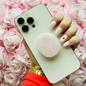May include: A light-colored smartphone with a pink pop-up grip is held against a backdrop of pink roses. The phone has three black camera lenses. The nails are painted light pink with a red heart on one nail. Gold bracelets and a red sleeve are visible.