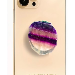 May include: A gold-colored smartphone with a triple-lens camera system and the text CRYSTALFY printed on the back. Attached to the center is a circular, polished gemstone accessory featuring horizontal bands of deep purple, white, and translucent hues.