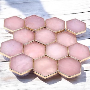 May include: A collection of pink hexagonal phone grips with gold-colored trim. The grips are arranged in a honeycomb pattern, showcasing the rose quartz material. The light pink color and gold accents create an elegant and stylish look.