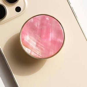 May include: A pink, circular phone grip with a gold-colored rim is attached to a gold-colored phone. The phone grip has a pearlescent finish. The word "CRYSTALFY" is printed on the phone.
