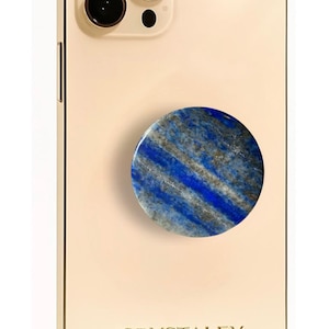 May include: A gold-colored smartphone with a triple-lens camera system and the text CRYSTALFY printed on the back. Attached to the center is a circular phone grip featuring a polished blue lapis lazuli stone with grey and white mineral inclusions.