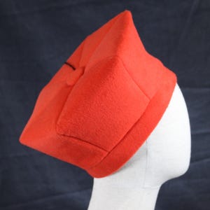 Peut inclure: Un chapeau rouge vif de forme carrée, avec une surface légèrement texturée. Le chapeau est présenté sur une tête de mannequin blanche, sur fond bleu foncé. Le chapeau présente un dessus plié et un bord bien défini.