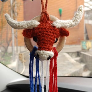 May include: A handmade car charm featuring a crocheted bull's head with white horns and a red body. The charm is adorned with blue, white, and red tassels, and hangs from a brown cord. The charm is hanging from a car's rearview mirror.