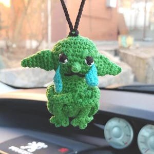 May include: A green crocheted figure with large ears, black eyes, and blue teardrops, hanging from a black string. The figure has a small black mustache and is suspended in front of a car dashboard.