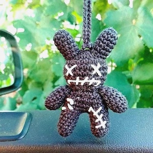 May include: A dark gray crocheted plush rabbit hanging ornament featuring white stitched X-shaped eyes and a stitched mouth. The rabbit has a textured yarn surface with white cross-stitch details on the chest and limbs, suspended by a matching cord.