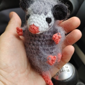 May include: A grey and white crocheted opossum plush toy with pink feet and a long tail. The toy is being held in a person's hand.