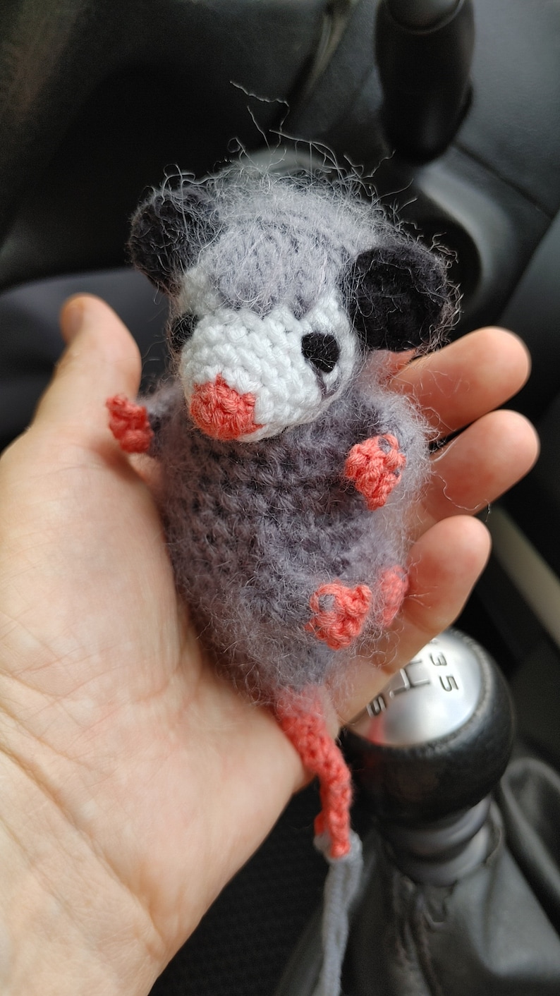 May include: A grey and white crocheted opossum plush toy with pink feet and a long tail. The toy is being held in a person's hand.