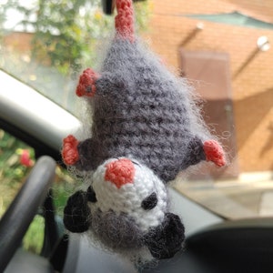 May include: A grey and white crocheted opossum hanging from a rear view mirror in a car. The opossum has pink ears and a pink nose.