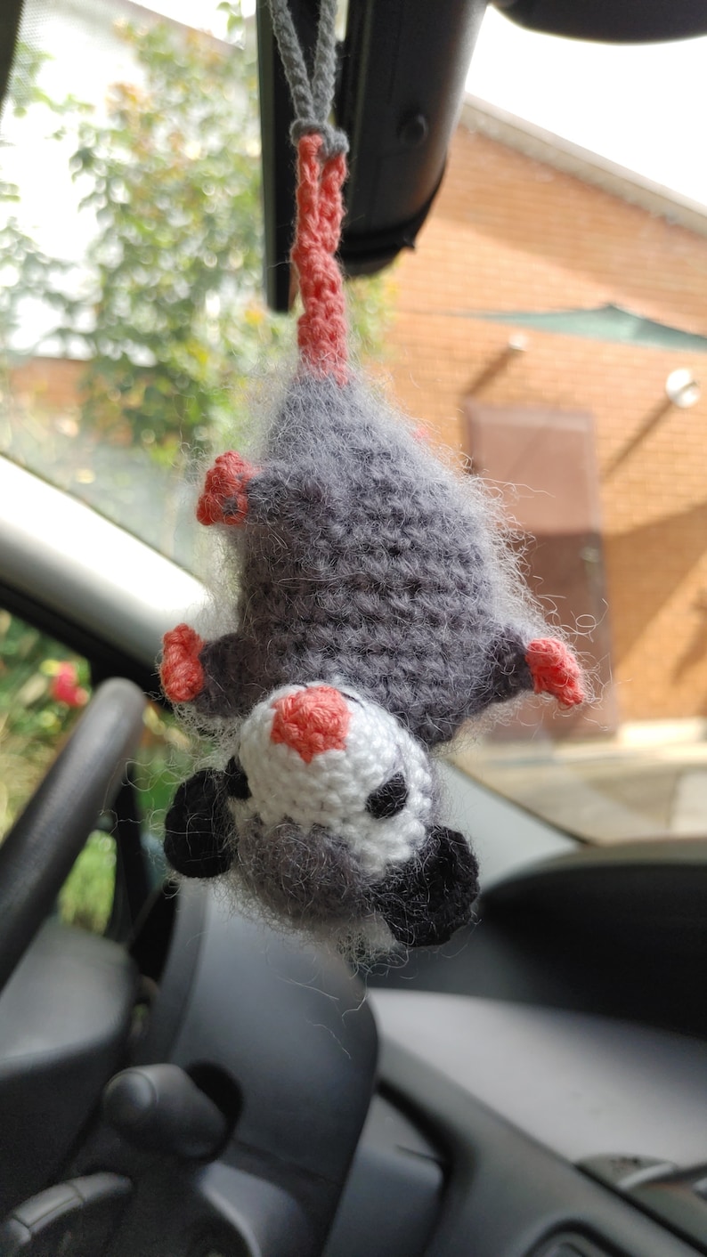 May include: A grey and white crocheted opossum hanging from a rear view mirror in a car. The opossum has pink ears and a pink nose.
