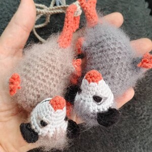 May include: Two crocheted opossum plushies, one grey and one beige, with orange feet and black eyes. The plushies are holding onto each other.