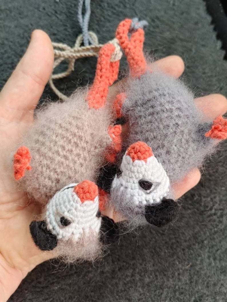 May include: Two crocheted opossum plushies, one grey and one beige, with orange feet and black eyes. The plushies are holding onto each other.