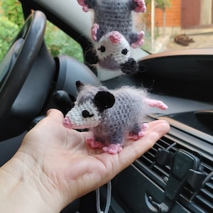 May include: Two crocheted opossum plushies, one hanging from a rearview mirror in a car and the other held in a hand. Both plushies are gray with pink accents and black eyes.