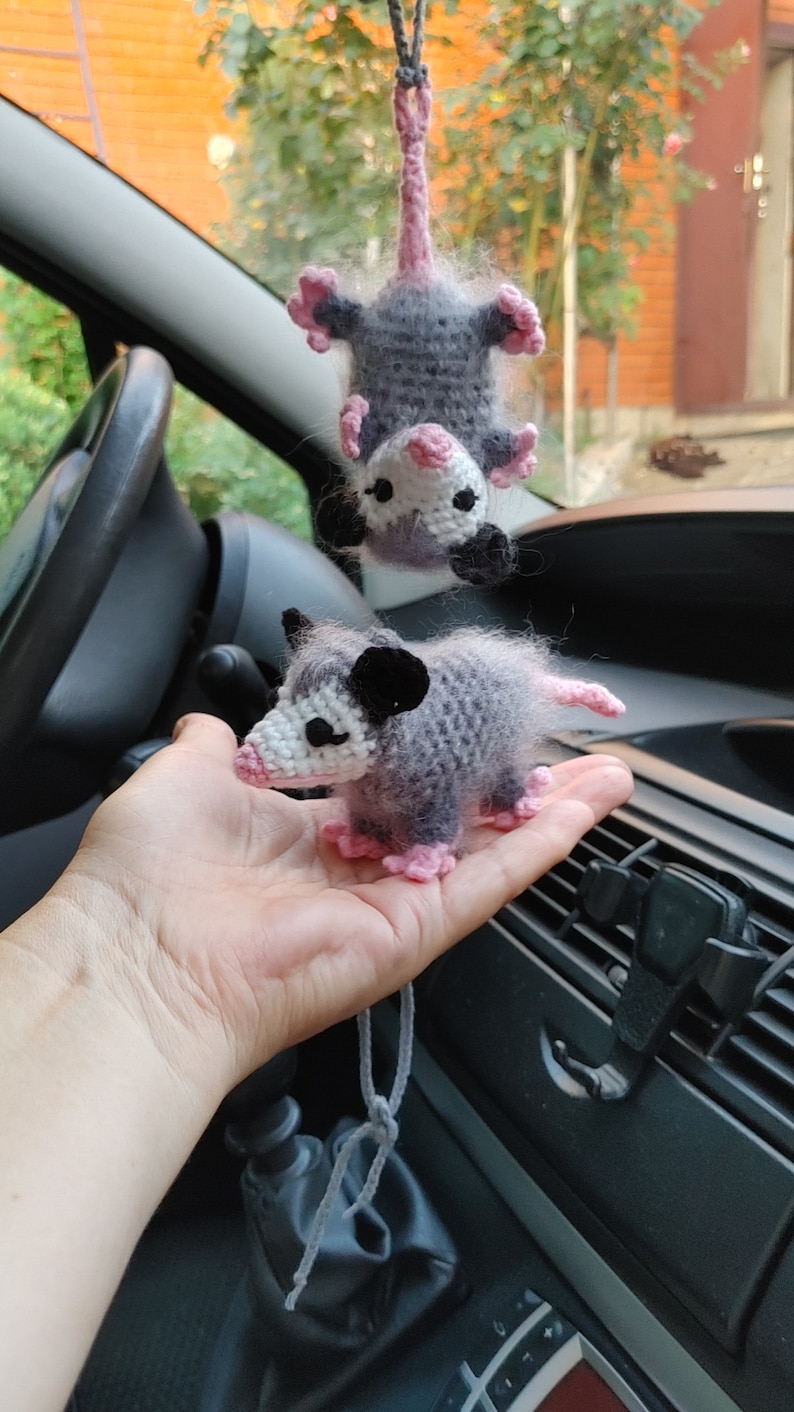 May include: Two crocheted opossum plushies, one hanging from a rearview mirror in a car and the other held in a hand. Both plushies are gray with pink accents and black eyes.