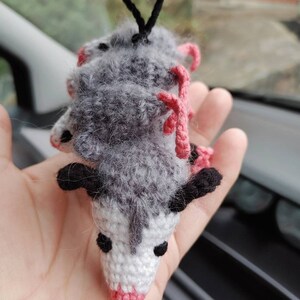 May include: A gray and white crocheted opossum hanging from a rearview mirror. The opossum has pink ears and a pink nose.