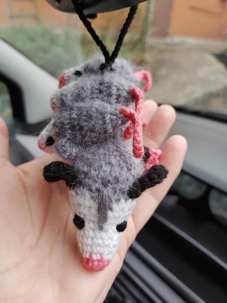 May include: A gray and white crocheted opossum hanging from a rearview mirror. The opossum has pink ears and a pink nose.