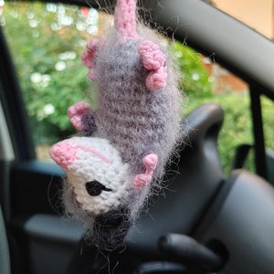 May include: A gray and white crocheted opossum hanging from a rearview mirror in a car. The opossum has pink ears and a pink tail.