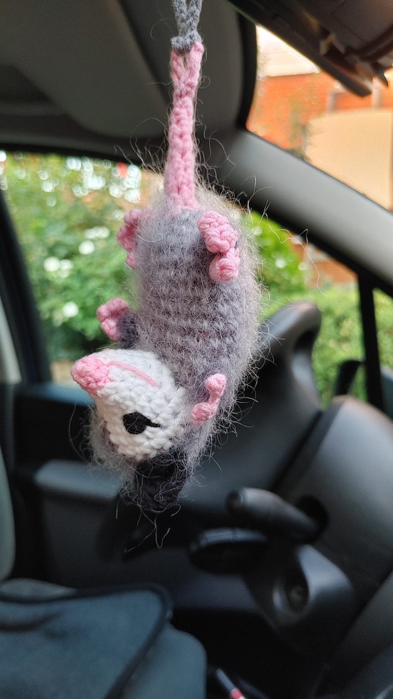 May include: A gray and white crocheted opossum hanging from a rearview mirror in a car. The opossum has pink ears and a pink tail.