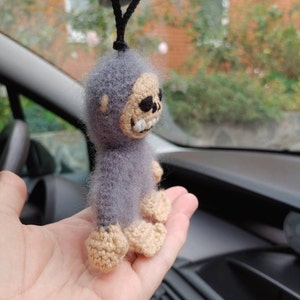 Puede incluir: Un juguete de peluche gris tejido a crochet con una cara de calavera y una cuerda negra que cuelga de la parte superior. El juguete est&aacute; siendo sostenido en la mano de una persona.