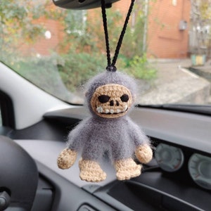 Puede incluir: Un mono de crochet gris con una cara de calavera colgando de un espejo retrovisor. El mono tiene un cuerpo de crochet beige y una cara de calavera de crochet blanco.