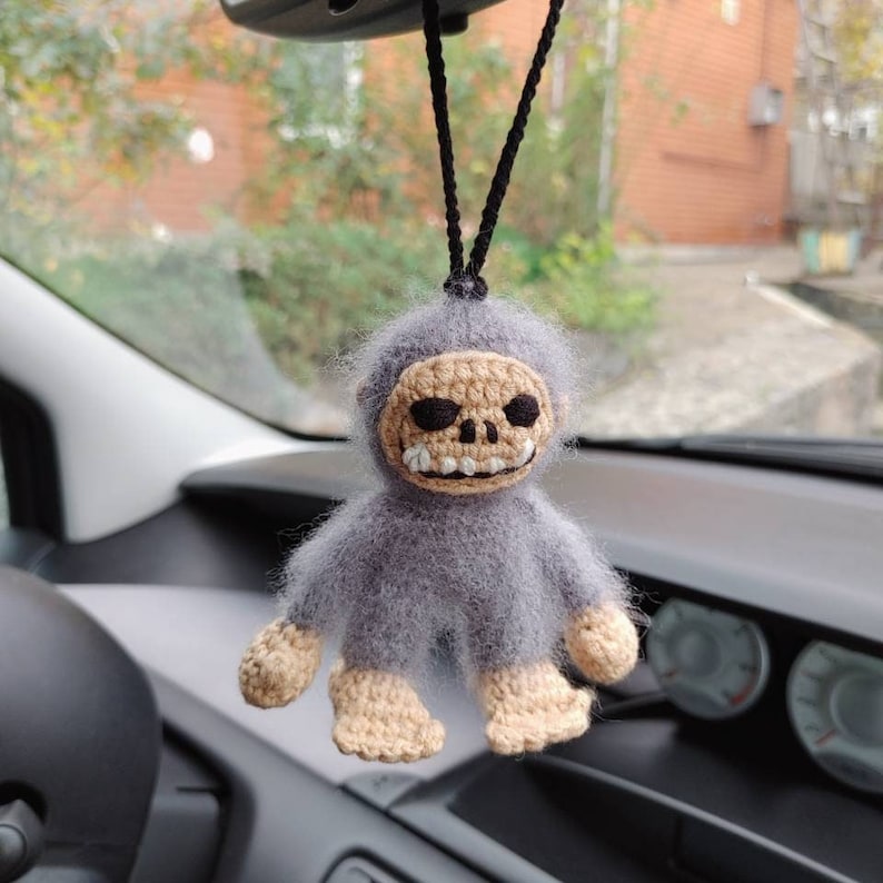 Puede incluir: Un mono de crochet gris con una cara de calavera colgando de un espejo retrovisor. El mono tiene un cuerpo de crochet beige y una cara de calavera de crochet blanco.