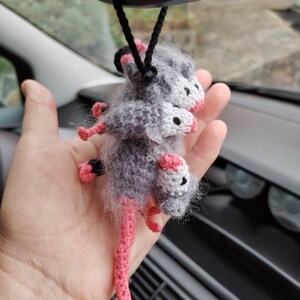 May include: A gray and white crocheted opossum with pink legs hanging from a rearview mirror in a car.