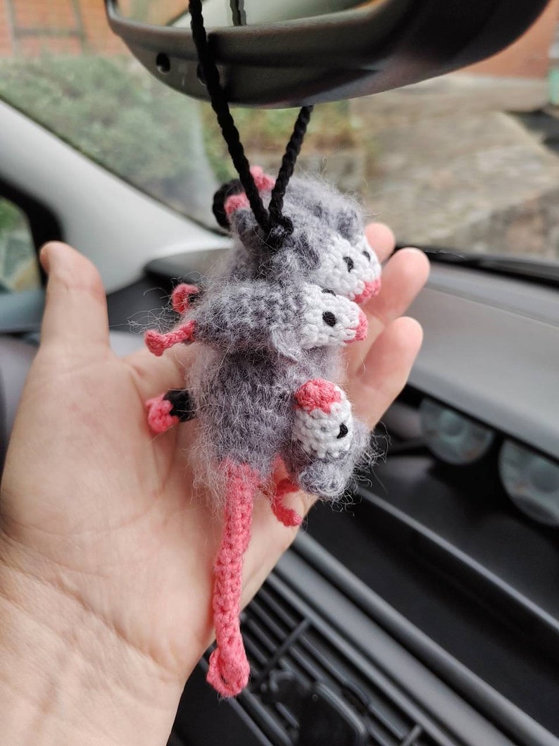 May include: A gray and white crocheted opossum with pink legs hanging from a rearview mirror in a car.