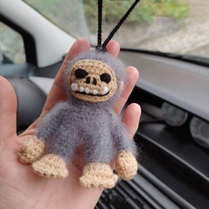 Puede incluir: Un juguete de peluche gris tejido a crochet con forma de calavera y una sonrisa de dientes. El juguete est&aacute; colgado de un espejo retrovisor en un coche.