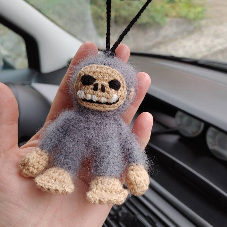Puede incluir: Un juguete de peluche gris tejido a crochet con forma de calavera y una sonrisa de dientes. El juguete est&aacute; colgado de un espejo retrovisor en un coche.