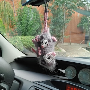 Könnte beinhalten: Gehäkelter possum-Anhänger für das Auto. Der Anhänger ist grau mit rosa Akzenten an Pfoten und Ohren. Das possum hat ein weißes Gesicht mit schwarzen Augen und einer rosa Nase. Aufgehängt an einer schwarzen Schnur.