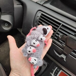 May include: A crocheted gray and white opossum with pink ears and nose. The opossum is curled up in a ball with its tail wrapped around its body.