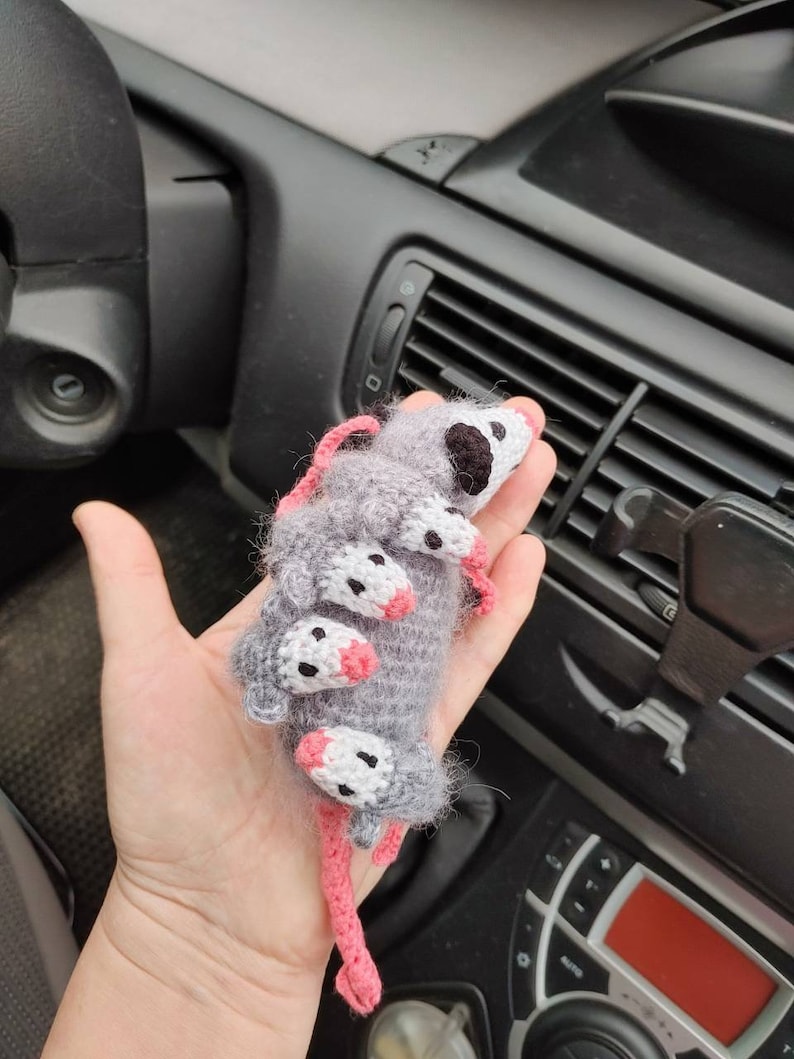 May include: A crocheted gray and white opossum with pink ears and nose. The opossum is curled up in a ball with its tail wrapped around its body.